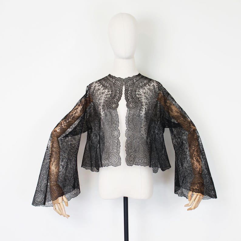 May include: A black lace jacket with wide, flared sleeves. The open-front jacket is displayed on a mannequin against a white background. The intricate lace design is visible throughout the garment, creating a delicate and elegant look.
