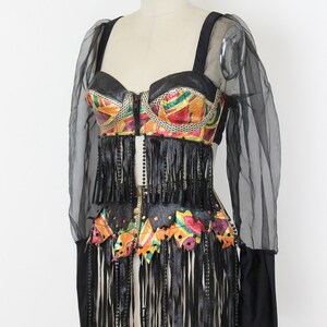 May include: A vintage, colorful, and embellished top and skirt set. The top features a black leather-like bodice with a zipper and colorful geometric patterns. The skirt has long black fringe. Sheer black sleeves complete the look.