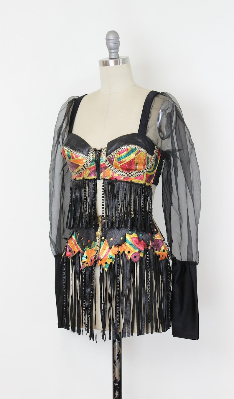 May include: A vintage, colorful, and embellished top and skirt set. The top features a black leather-like bodice with a zipper and colorful geometric patterns. The skirt has long black fringe. Sheer black sleeves complete the look.