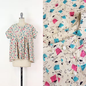 May include: A cream-coloured short-sleeved blouse featuring a pattern of pink and teal geometric shapes. The blouse has a round collar and a button-up front. The fabric is lightweight, suggesting a vintage style.