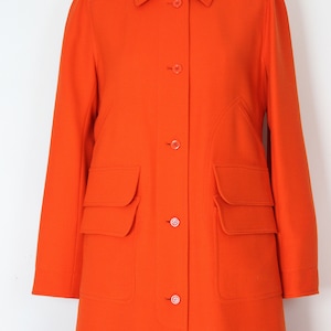 May include: A long, bright orange coat with a collared neckline and button closure. The coat has two large flap pockets on the front and a fitted silhouette.
