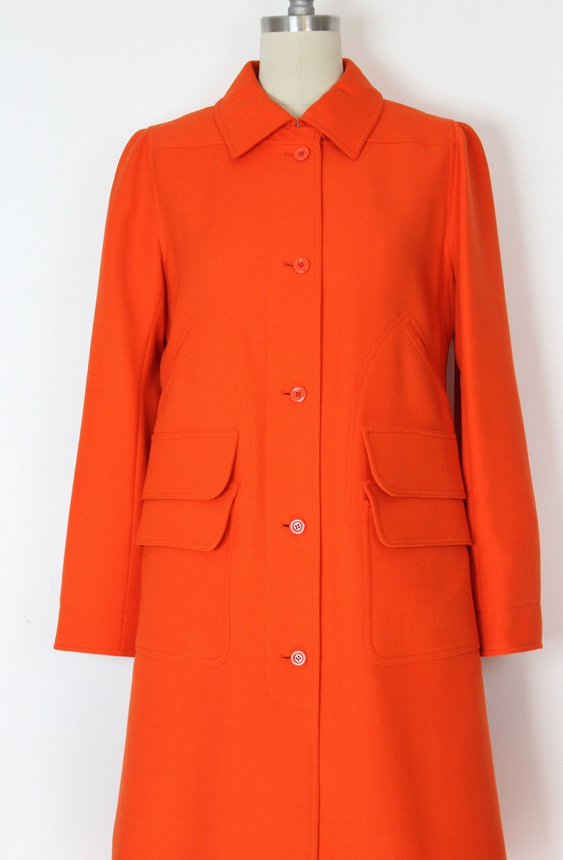 May include: A long, bright orange coat with a collared neckline and button closure. The coat has two large flap pockets on the front and a fitted silhouette.