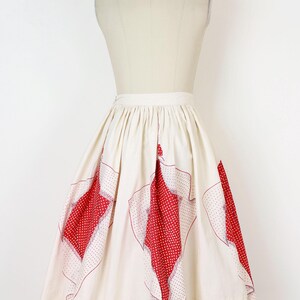 May include: Cream-colored vintage skirt with red polka-dot and patterned accents. The skirt is displayed on a mannequin, showcasing its flared shape and unique design. A classic piece for a retro-inspired wardrobe.