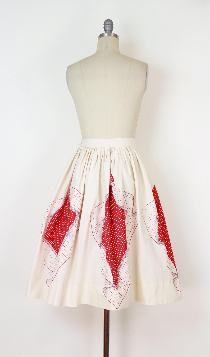May include: Cream-colored vintage skirt with red polka-dot and patterned accents. The skirt is displayed on a mannequin, showcasing its flared shape and unique design. A classic piece for a retro-inspired wardrobe.