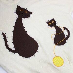 May include: A white knitted sweater with two brown embroidered cats. The larger cat is sitting with its tail curled around its body. The smaller cat is playing with a yellow ball of yarn.