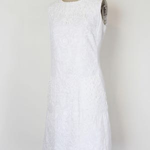 May include: White sleeveless dress with a round neckline and a subtle floral pattern. The dress features delicate lace detailing around the neckline and on the side panels. The dress is displayed on a mannequin.