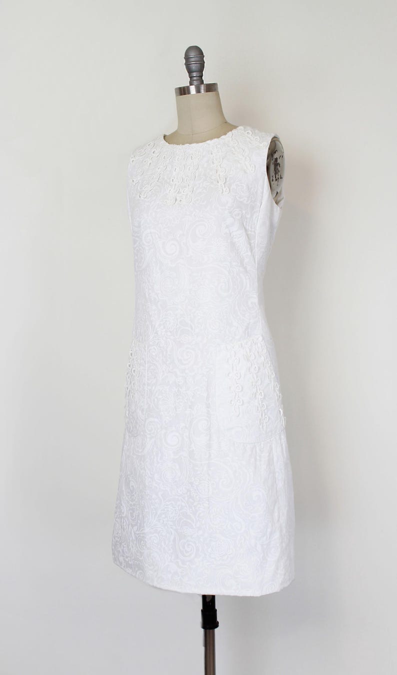 May include: White sleeveless dress with a round neckline and a subtle floral pattern. The dress features delicate lace detailing around the neckline and on the side panels. The dress is displayed on a mannequin.