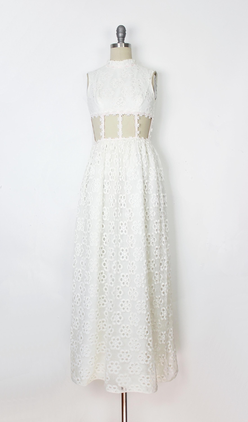 May include: White sleeveless maxi dress with a high neckline and floral eyelet pattern. The dress features a sheer panel at the waist. The dress is displayed on a mannequin.