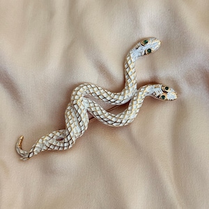 Puede incluir: Un broche de serpiente de dos cabezas con escamas de esmalte blanco y contornos dorados. Cada cabeza de serpiente tiene dos pequeñas piedras verdes incrustadas como ojos. El cuerpo metálico presenta un patrón de diamantes.