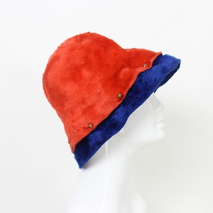 Puede incluir: Un sombrero de cubo rojo y azul con una textura peluda y detalles dorados. El sombrero está hecho de dos telas diferentes, una peluda y otra lisa.