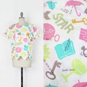 May include: A white, short-sleeved top with a unique scalloped hem. The fabric is patterned with colorful illustrations of keys, stars, umbrellas, and text. The word "STAR" is prominently displayed. The top is displayed on a mannequin.