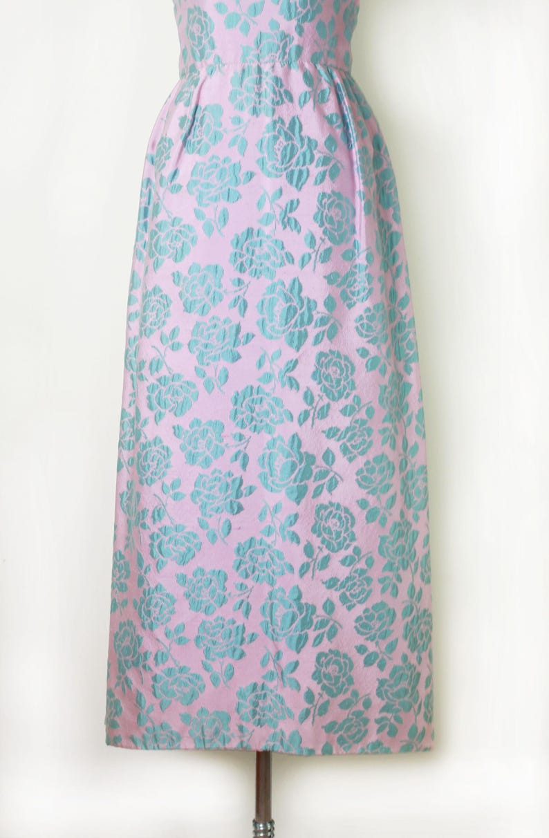 May include: A vintage-style dress with a pink background and a repeating pattern of teal roses. The dress has a fitted bodice and a flared skirt, ideal for a formal event or special occasion. The fabric has a subtle sheen.
