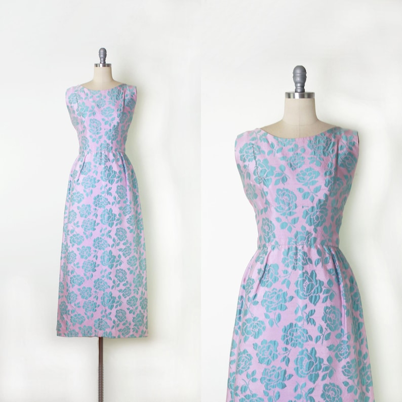 May include: A vintage, sleeveless dress in a light pink hue, adorned with a repeating pattern of teal-colored floral designs. The dress features a fitted bodice, a defined waist, and a flowing skirt, displayed on a dress form.