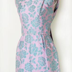 May include: A sleeveless, vintage-style dress in a pink fabric with a teal floral pattern. The dress features a fitted bodice, a defined waist, and a knee-length skirt. The floral design is a repeating pattern of roses.