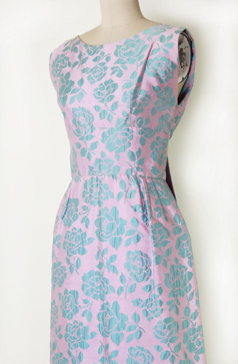 May include: A sleeveless, vintage-style dress in a pink fabric with a teal floral pattern. The dress features a fitted bodice, a defined waist, and a knee-length skirt. The floral design is a repeating pattern of roses.
