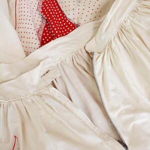 May include: Cream-colored garment with a zipper, possibly a skirt or dress, featuring gathered fabric details. A red and white polka-dot pattern is visible in the background, along with a red and white patterned accent. The overall aesthetic suggests vintage or retro clothing.