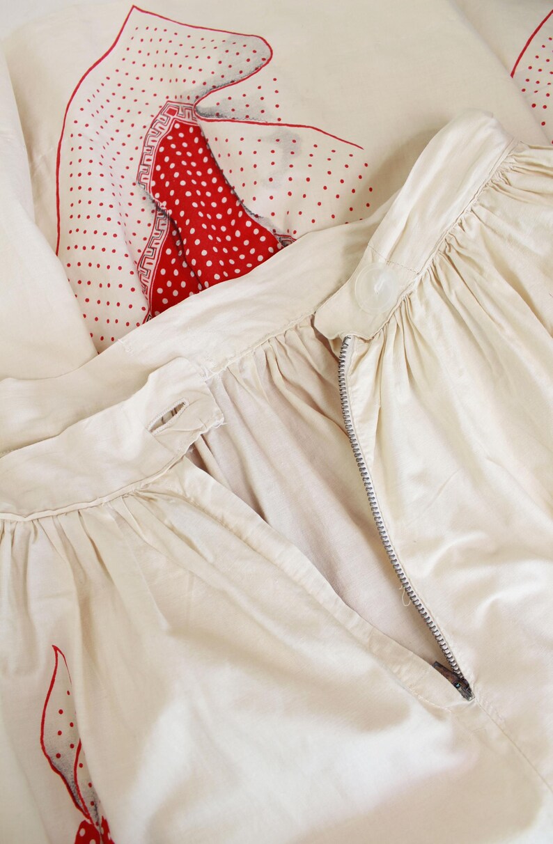 May include: Cream-colored garment with a zipper, possibly a skirt or dress, featuring gathered fabric details. A red and white polka-dot pattern is visible in the background, along with a red and white patterned accent. The overall aesthetic suggests vintage or retro clothing.