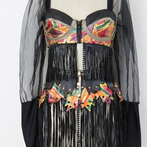May include: A black and colorful bustier-style top with fringe and beaded accents. The top features a colorful, patterned design on the bodice and waist, with long black fringe and black sleeves.