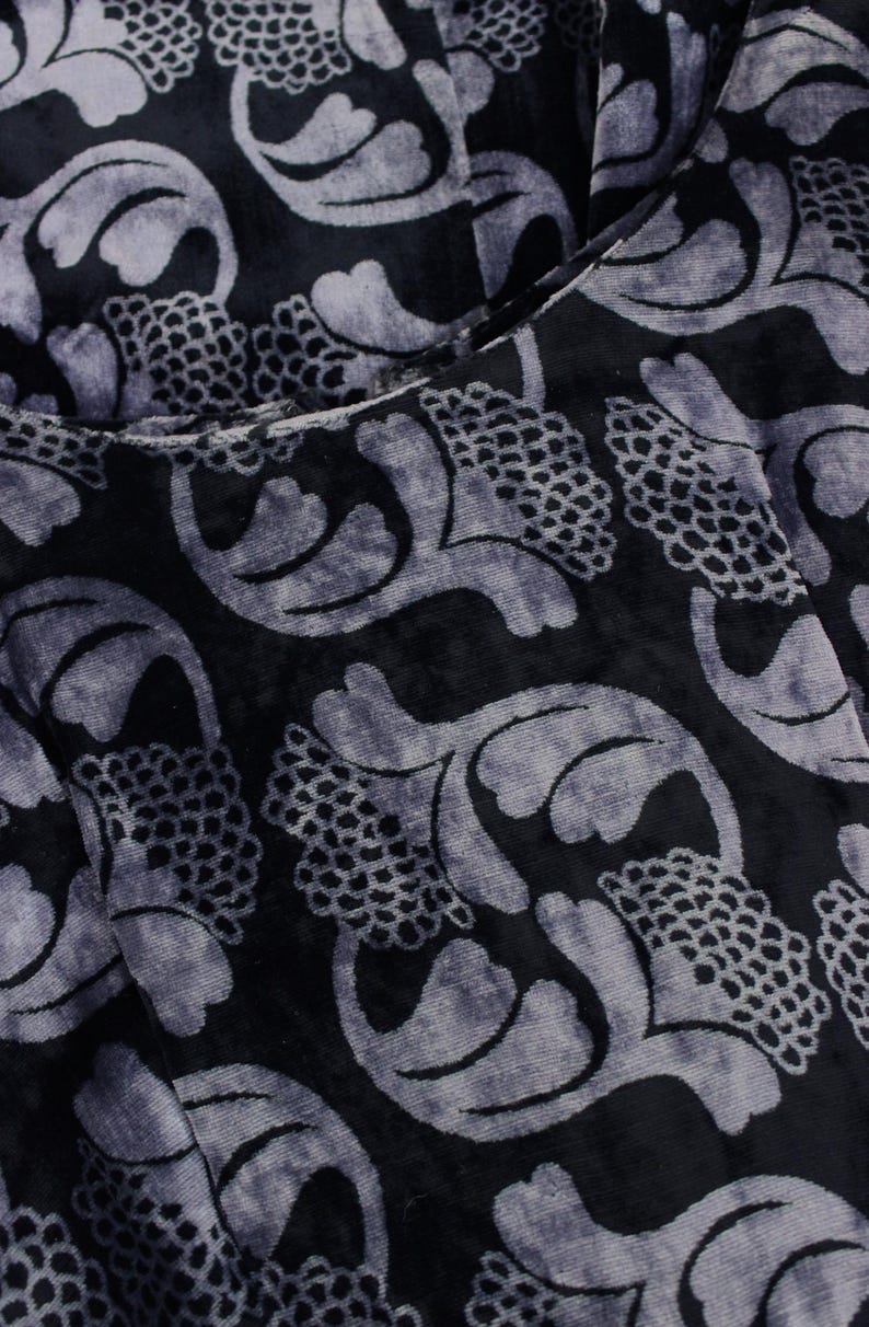 May include: Close-up of a dark fabric with a velvet texture. The fabric features a repeating pattern of stylized floral and leaf shapes in a lighter shade of gray, creating a contrast against the dark background. The design has a vintage or art nouveau feel.
