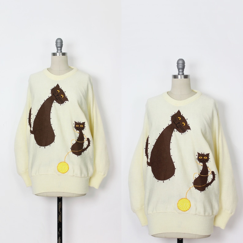 May include: Two cream-colored sweaters with brown embroidered cats and a ball of yarn. The cats are stylized and have yellow eyes.