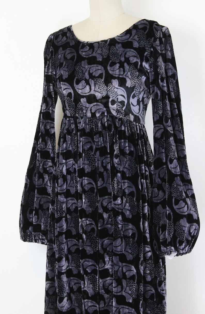 May include: A long black velvet dress with a gray floral pattern. The dress features long, voluminous sleeves and a gathered waist. The fabric has a soft, textured appearance, and the dress is displayed on a mannequin.