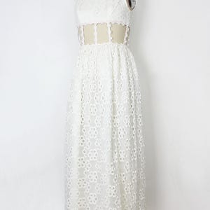 May include: A white, sleeveless, floor-length dress on a mannequin. The dress has a high neckline and a floral lace pattern. The waist has a sheer panel with delicate trim. The dress is made of a lightweight fabric.