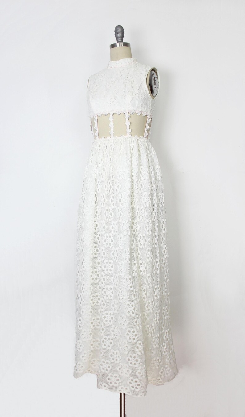 May include: A white, sleeveless, floor-length dress on a mannequin. The dress has a high neckline and a floral lace pattern. The waist has a sheer panel with delicate trim. The dress is made of a lightweight fabric.