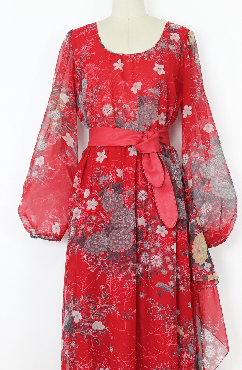 May include: A red maxi dress with a floral print and a matching red sash. The dress has long sleeves and a flowing skirt.