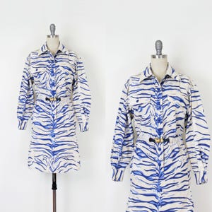 Puede incluir: Vestido de manga larga con estampado de rayas de cebra azules y blancas. El vestido tiene cuello camisero, cierre de botones en la parte delantera y cinturón negro en la cintura. El vestido se exhibe en un maniquí.