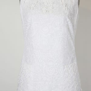 May include: White sleeveless dress with a floral pattern and decorative eyelet detailing. The dress features a round neckline with embroidered accents and two front pockets. The fabric has a subtle, all-over embossed design.