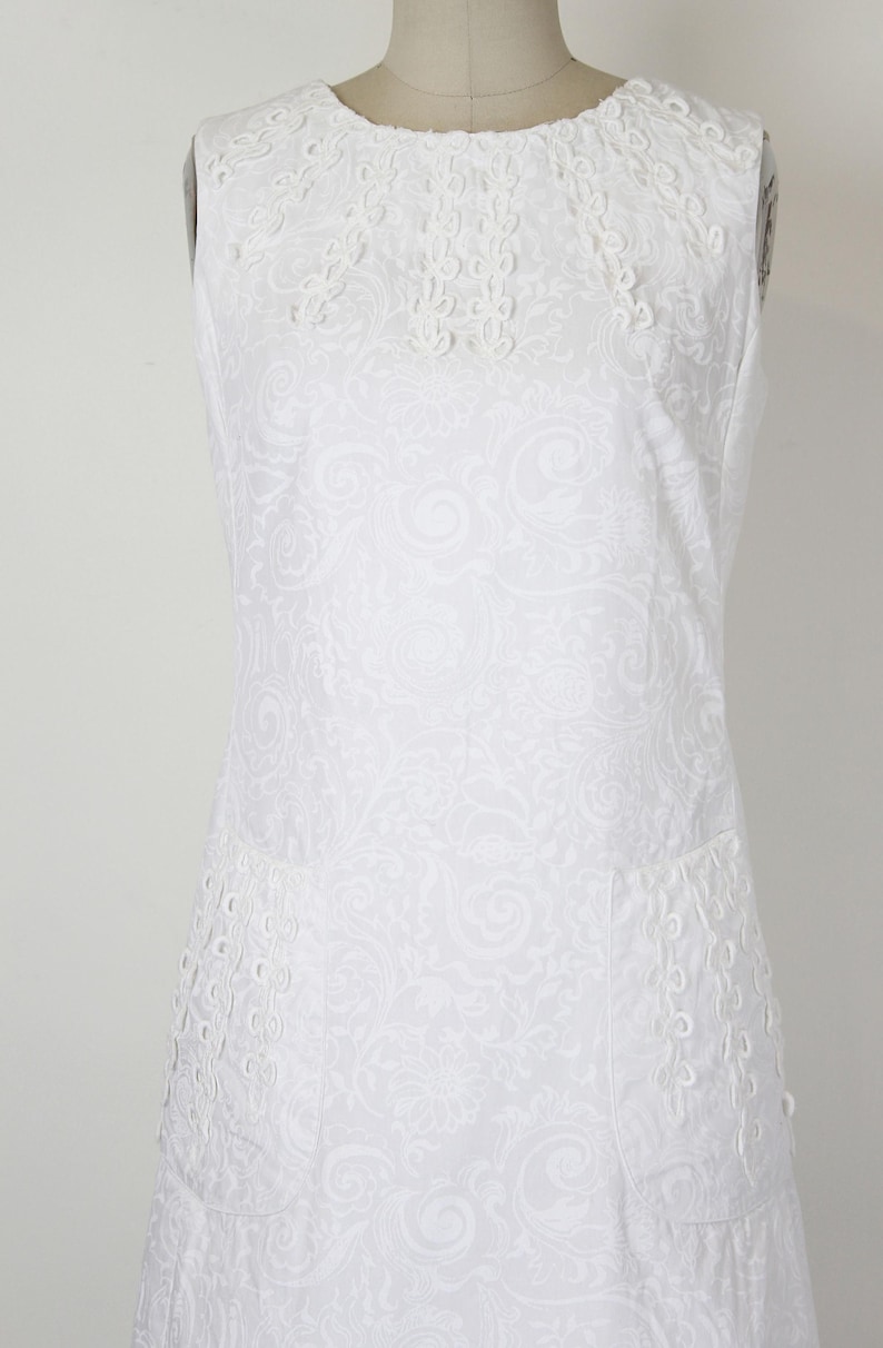 May include: White sleeveless dress with a floral pattern and decorative eyelet detailing. The dress features a round neckline with embroidered accents and two front pockets. The fabric has a subtle, all-over embossed design.
