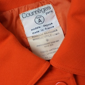 May include: Orange jacket with a white label that reads "Courr&egrave;ges" and "Taille A" in a circle. The label also says "modele depose made in france" and "FABRIQUE EN FRANCE MODELE/STYLE: 17801". The label also includes care instructions: "PROFESSIONNALLY CLEAN ONLY WARM IRON" and "TISSUS-MATERIAL 95% LAINEWOON 5% Pol".