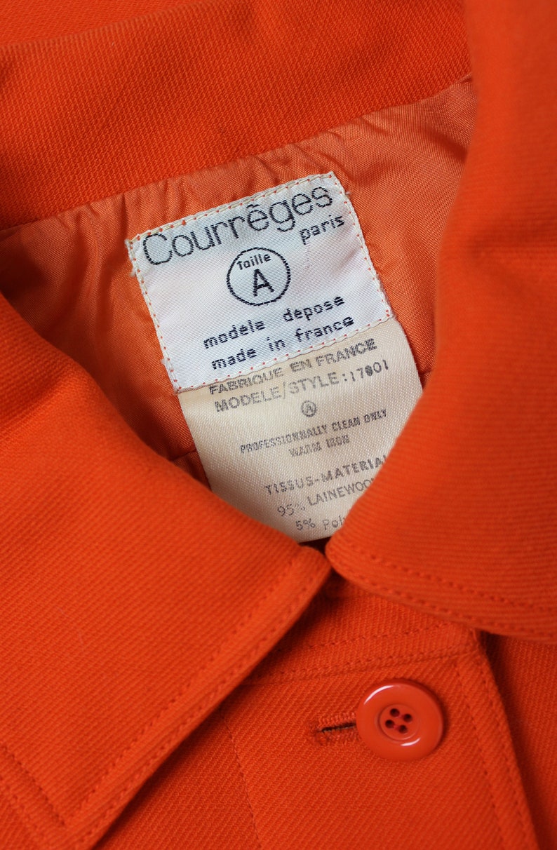 May include: Orange jacket with a white label that reads "Courr&egrave;ges" and "Taille A" in a circle. The label also says "modele depose made in france" and "FABRIQUE EN FRANCE MODELE/STYLE: 17801". The label also includes care instructions: "PROFESSIONNALLY CLEAN ONLY WARM IRON" and "TISSUS-MATERIAL 95% LAINEWOON 5% Pol".