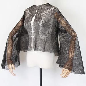 May include: Black lace jacket with long, flared sleeves. The jacket is displayed on a mannequin, showcasing its intricate floral and paisley patterns. The open front design and delicate fabric create an elegant, vintage aesthetic.