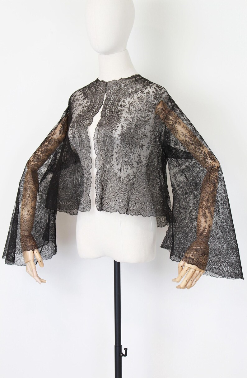 May include: Black lace jacket with long, flared sleeves. The jacket is displayed on a mannequin, showcasing its intricate floral and paisley patterns. The open front design and delicate fabric create an elegant, vintage aesthetic.