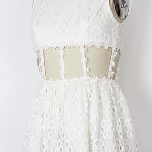 May include: A white lace dress with a high neckline and a fitted bodice. The dress features a floral lace pattern and a sheer panel at the waist, trimmed with small pink flowers. The skirt is flared, creating a romantic silhouette.