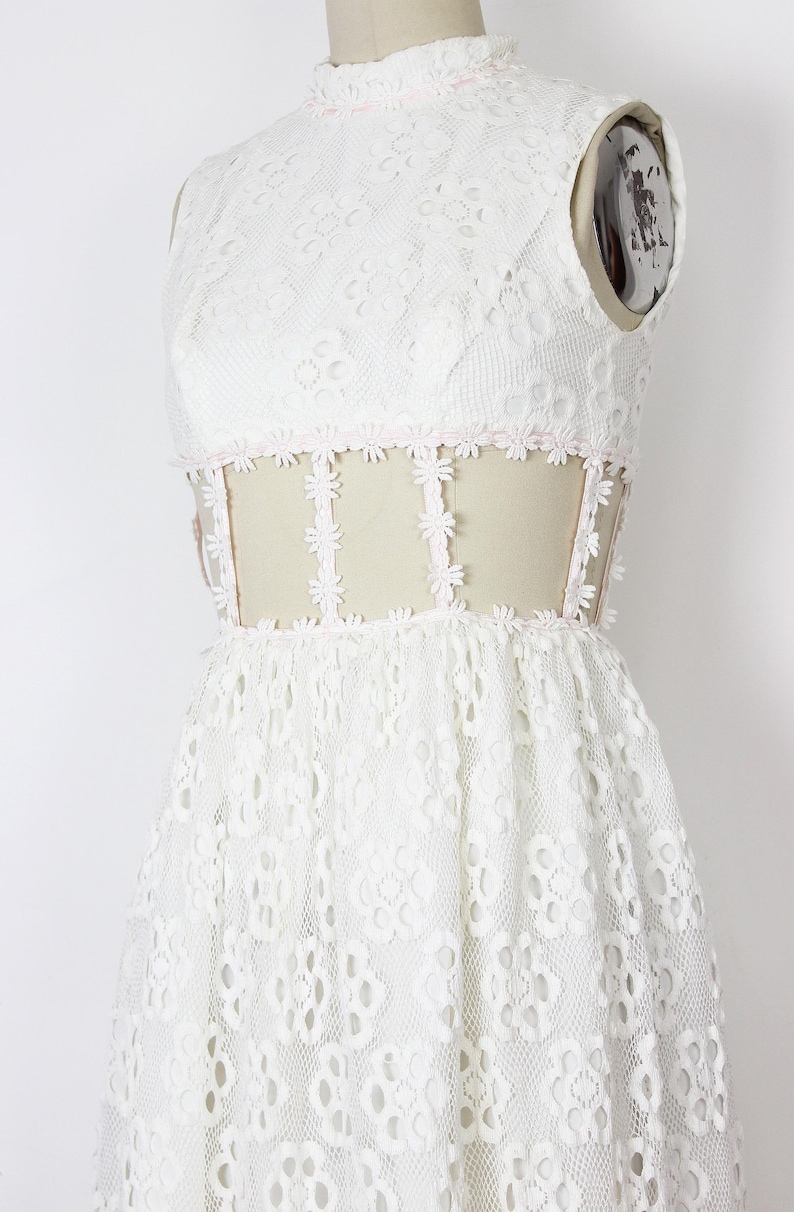 May include: A white lace dress with a high neckline and a fitted bodice. The dress features a floral lace pattern and a sheer panel at the waist, trimmed with small pink flowers. The skirt is flared, creating a romantic silhouette.