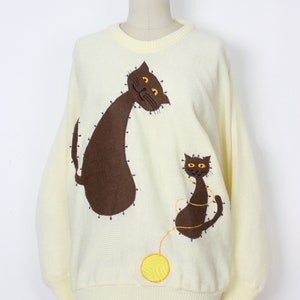 May include: A pale yellow knitted sweater with a brown cat and a smaller brown kitten embroidered on the front. The cat is looking to the left and the kitten is looking to the right. A yellow yarn ball is embroidered below the kitten.