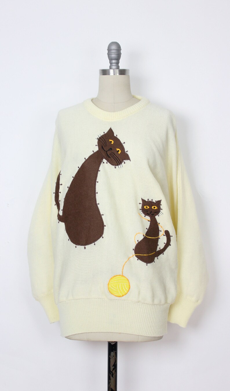 May include: A pale yellow knitted sweater with a brown cat and a smaller brown kitten embroidered on the front. The cat is looking to the left and the kitten is looking to the right. A yellow yarn ball is embroidered below the kitten.