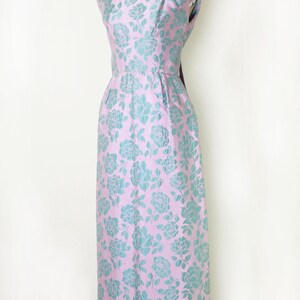 May include: A sleeveless, floor-length dress in a light pink fabric with a repeating pattern of teal-colored floral designs. The dress is displayed on a mannequin, showcasing its fitted bodice and flared skirt. The dress has a scoop neckline.