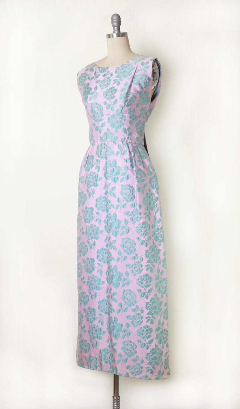 May include: A sleeveless, floor-length dress in a light pink fabric with a repeating pattern of teal-colored floral designs. The dress is displayed on a mannequin, showcasing its fitted bodice and flared skirt. The dress has a scoop neckline.