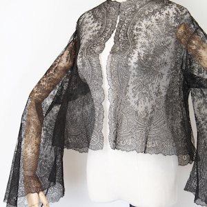 May include: Black lace jacket with a floral pattern and flared sleeves. The jacket is sheer and has a scalloped edge. The garment is displayed on a mannequin against a white background. A vintage-inspired piece.