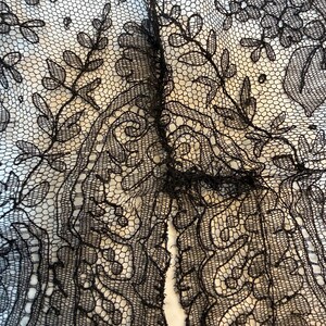 May include: Close-up of black lace fabric with floral and leaf patterns. The lace is intricately designed with delicate details, creating a textured appearance. The background is a light color, providing contrast to the dark lace.
