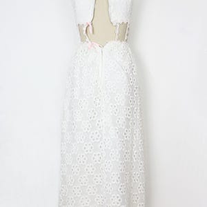 May include: White eyelet dress with a high neckline and open back. The dress features a floral pattern and is displayed on a mannequin. Pink bows accent the back and neckline. A vintage-inspired, elegant design.