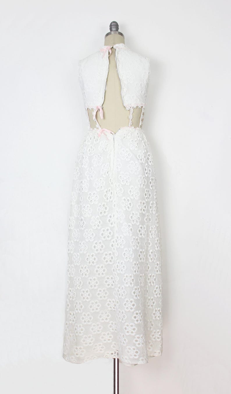 May include: White eyelet dress with a high neckline and open back. The dress features a floral pattern and is displayed on a mannequin. Pink bows accent the back and neckline. A vintage-inspired, elegant design.
