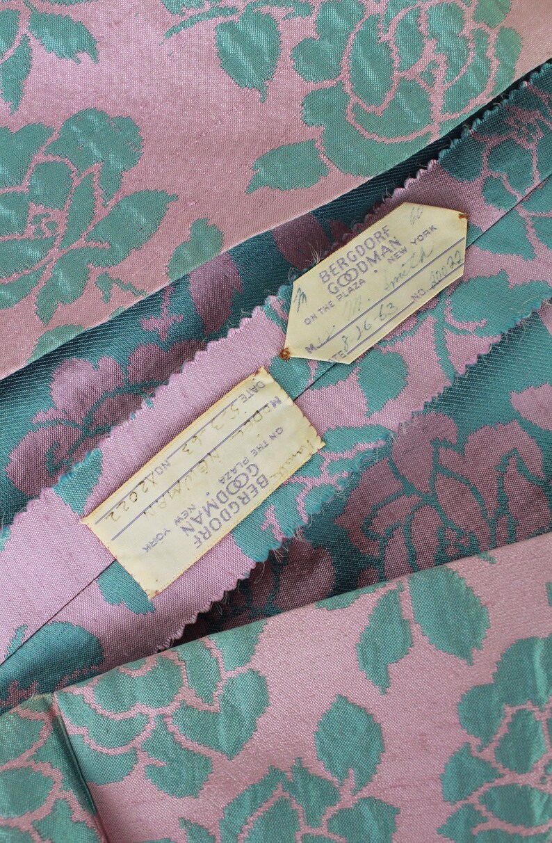 May include: Close-up of vintage fabric with a floral pattern in pink and teal. The fabric has a textured appearance. Two tags from Bergdorf Goodman, New York, are visible, with handwritten details including a date.