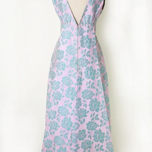 May include: A sleeveless, floor-length dress with a V-neck and a floral pattern. The dress is a light pink color with a teal floral design. The dress is displayed on a mannequin.