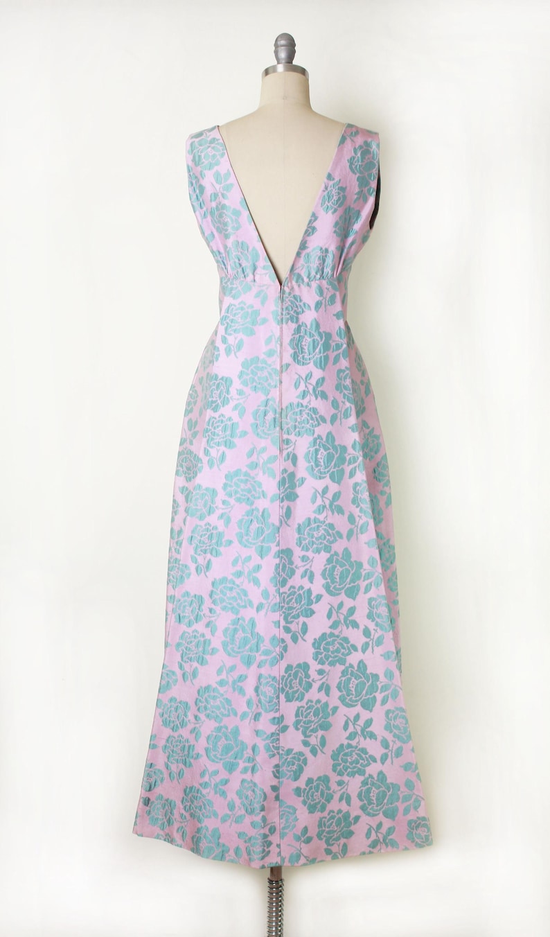 May include: A sleeveless, floor-length dress with a V-neck and a floral pattern. The dress is a light pink color with a teal floral design. The dress is displayed on a mannequin.