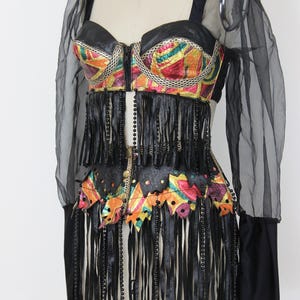 May include: A vintage-style bustier top with a colorful, patterned bodice and black leather fringe. The top features a zipper closure and is paired with sheer black sleeves and a black leather belt.