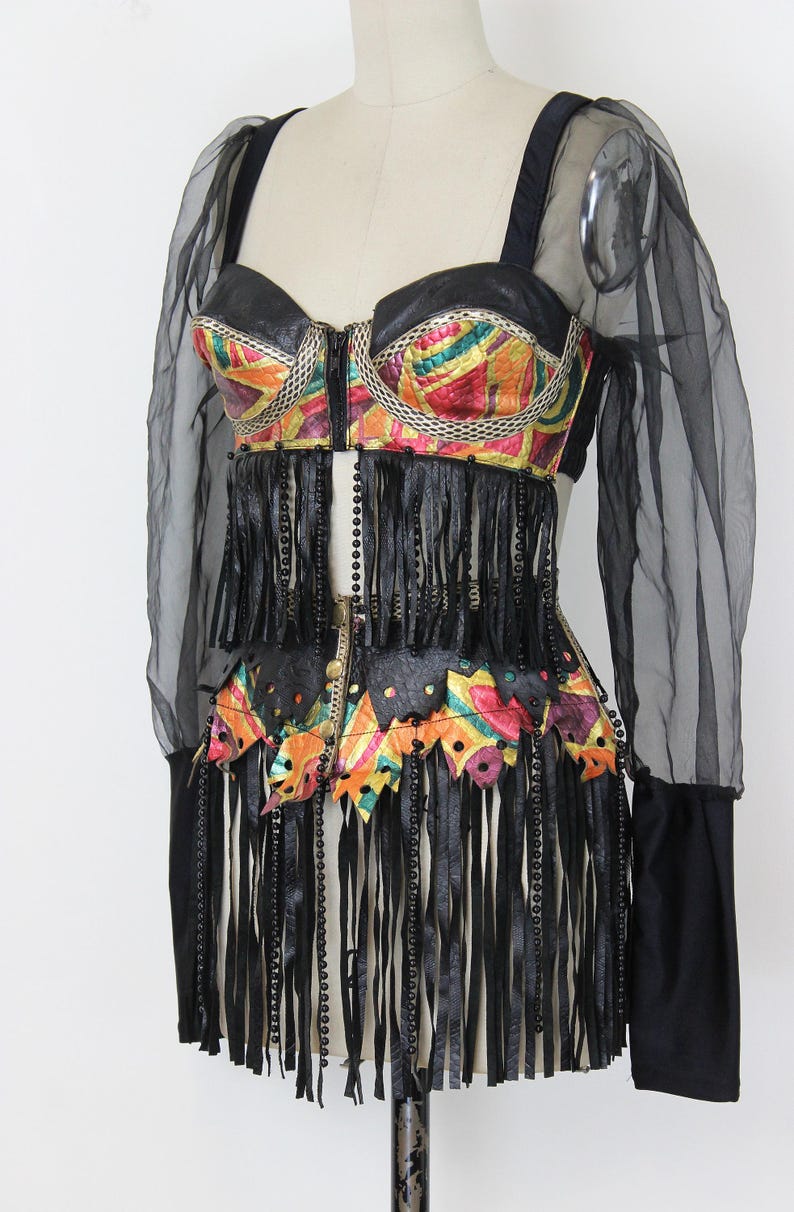 May include: A vintage-style bustier top with a colorful, patterned bodice and black leather fringe. The top features a zipper closure and is paired with sheer black sleeves and a black leather belt.